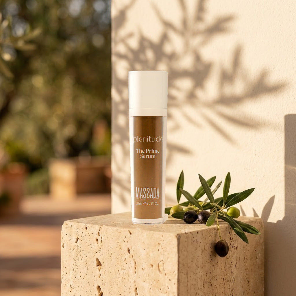 Massada Plenitude The Prime Serum 50 ml sobre un cubo de travertino, junto a una rama de hojas verdes con luz cálida